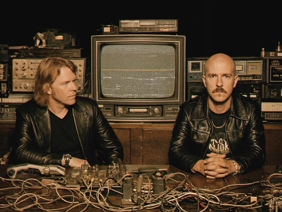 Two figures in conversation surrounded by vintage broadcast equipment, warm amber light, a monitor displaying waveforms between them